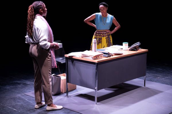 Production still of two female actors looking at each other next to a large desk