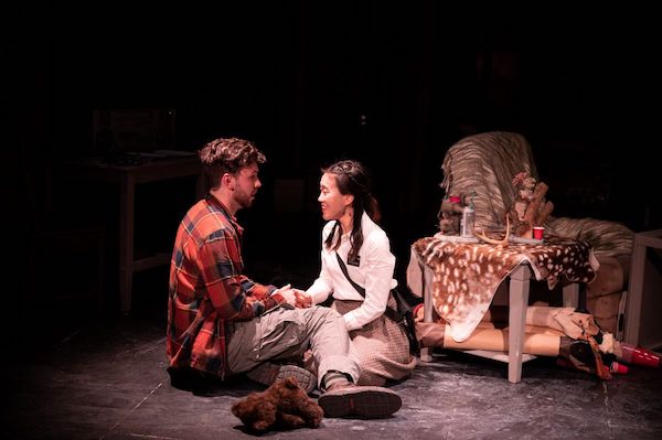 Production still of two actors sitting on the ground next to a dressed table and chair, holding hands