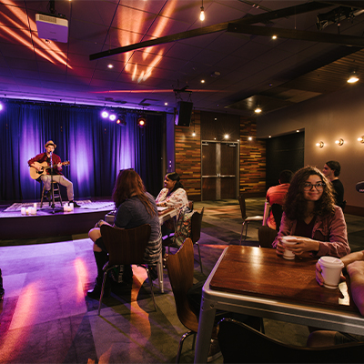 a guitar player on a stage in the background with students at tables watching him while drinking coffee in the foreground