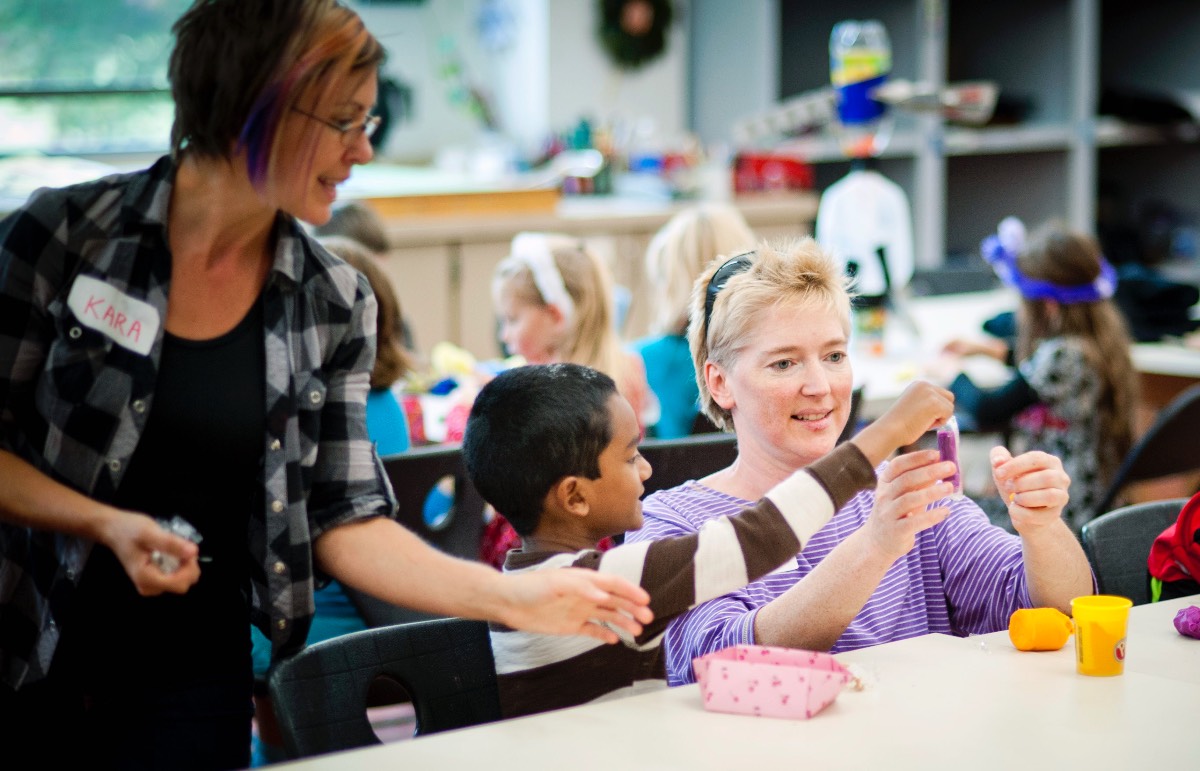 Children sit at long tables in a classroom creating pieces of art. In the foreground, one student is aided by two smiling adults.